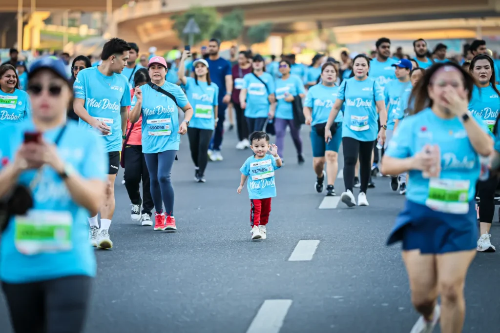 Участники забега в голубых футболках с надписью „Mai Dubai“, среди них — ребёнок, идущий по дороге