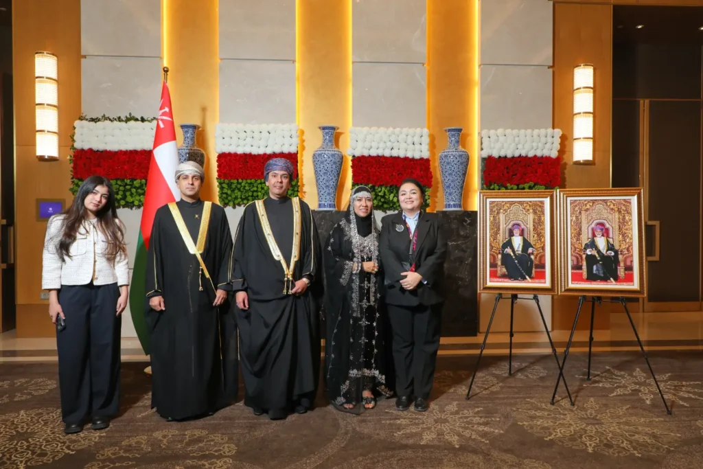 A group of people in traditional and formal attire posing at a ceremonial event with Omani flags and framed portraits in the background