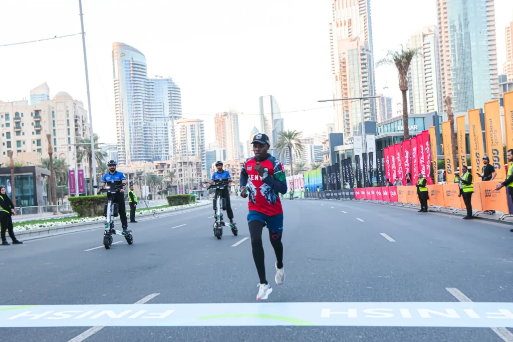 Бегун пересекает финишную линию на марафоне в Дубае, на заднем плане — сотрудники на электросамокатах и высотные здания