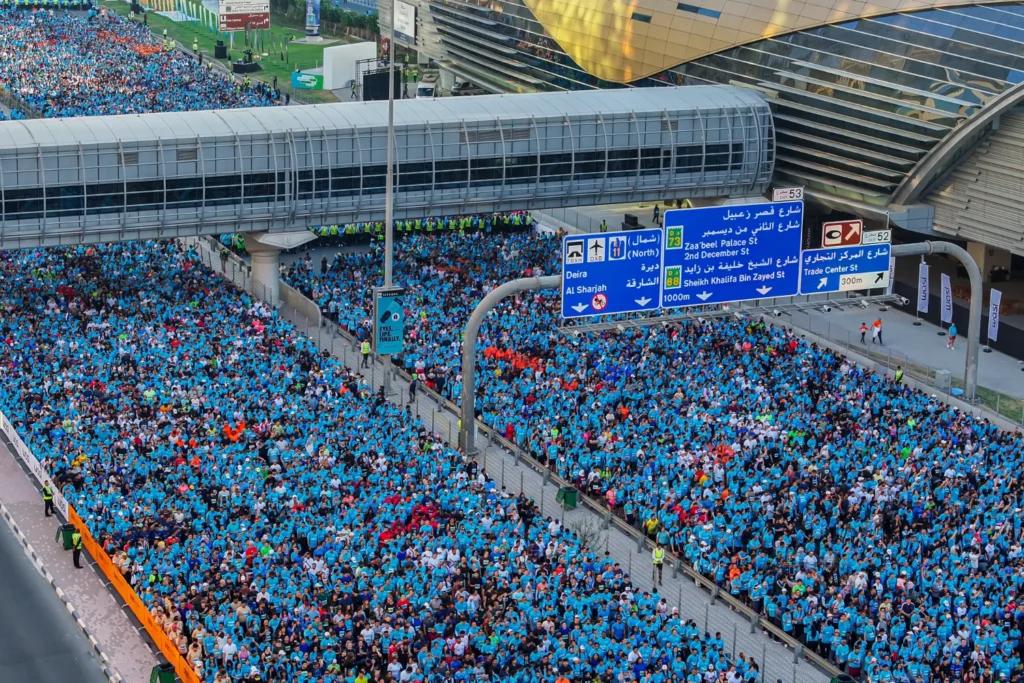 Участники Dubai Run заполняют трассу Sheikh Zayed Road; видны дорожные знаки с указанием направлений к Deira, Al Sharjah и другим районам Дубая