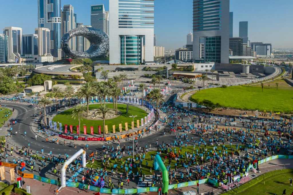 Спортивный фестиваль в Дубае: бегуны на городской трассе, на заднем плане — офисные здания и арт-объект в виде кольца