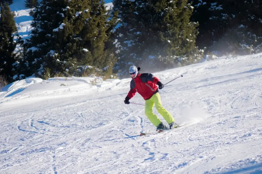 Лыжник в красной куртке и жёлтых брюках спускается по заснеженному склону, окружённому елями