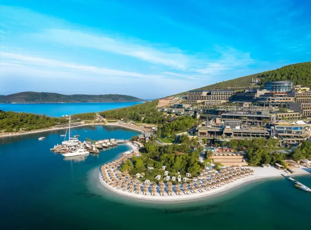 Панорамный вид на Lujo Hotel Bodrum с его частной бухтой, пляжем с лежаками, яхтами у пирса и зелёными холмами на фоне Эгейского моря