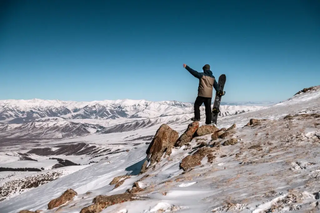Сноубордист на вершине горы, держит сноуборд, смотрит на заснеженные хребты
