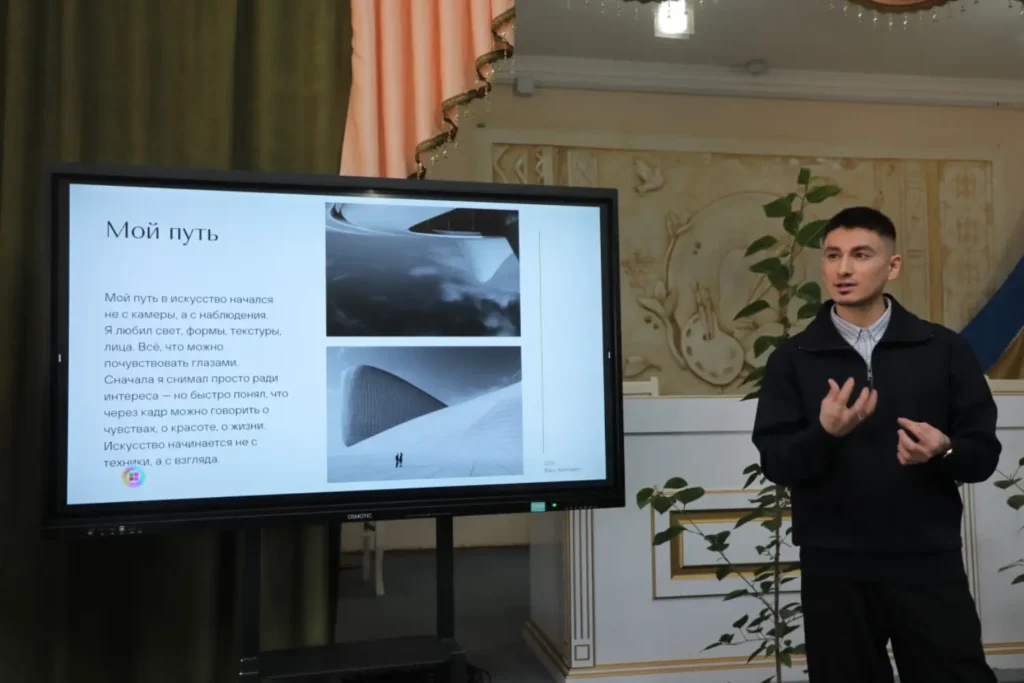 A person giving a presentation next to a large screen displaying text and photographs, speaking to an audience in an indoor setting