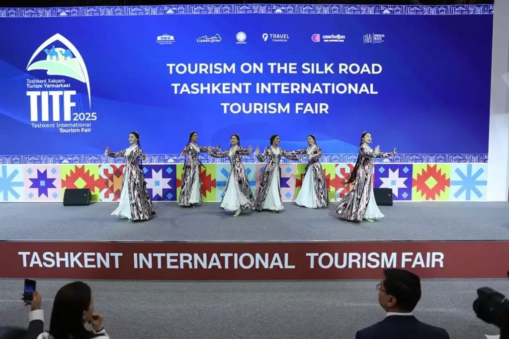 A group of six dancers in traditional Uzbek costumes performs a synchronized dance on the stage at the Tashkent International Tourism Fair (TITF 2025). In the background, there is a large screen displaying the event’s logo and the inscription «Tourism on the Silk Road».