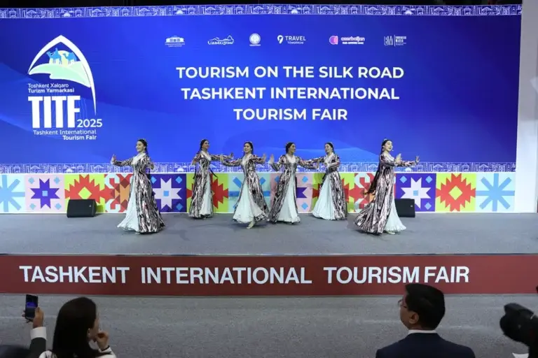 A group of six dancers in traditional Uzbek costumes performs a synchronized dance on the stage at the Tashkent International Tourism Fair (TITF 2025). In the background, there is a large screen displaying the event’s logo and the inscription «Tourism on the Silk Road».
