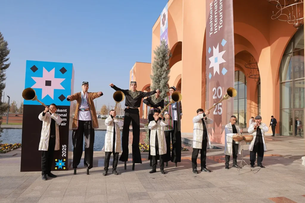 A group of artists in national costumes holding musical instruments stands in front of the Tashkent International Tourism Fair 2025 banner.