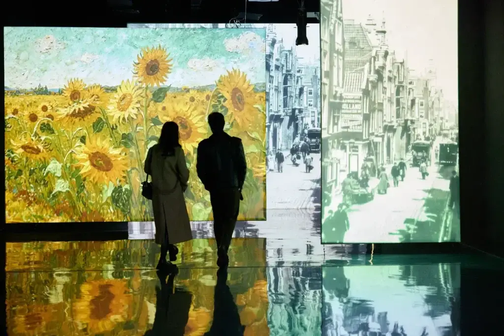 A couple of people stand in front of a multimedia exhibition: on the left is a bright painting with sunflowers by Van Gogh, on the right is a black-and-white urban landscape. The reflections of the paintings are visible on the glossy floor.