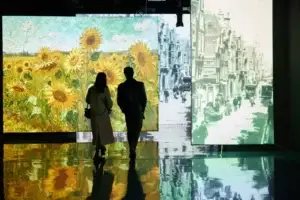 A couple of people stand in front of a multimedia exhibition: on the left is a bright painting with sunflowers by Van Gogh, on the right is a black-and-white urban landscape. The reflections of the paintings are visible on the glossy floor.