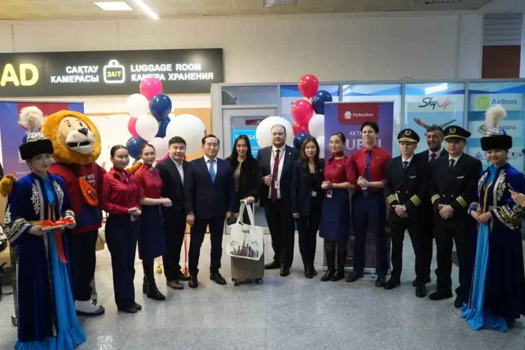 A group of people at the airport: pilots, airline staff in red uniforms, performers in traditional costumes, and a lion mascot in front of the «Luggage Room» sign and balloons