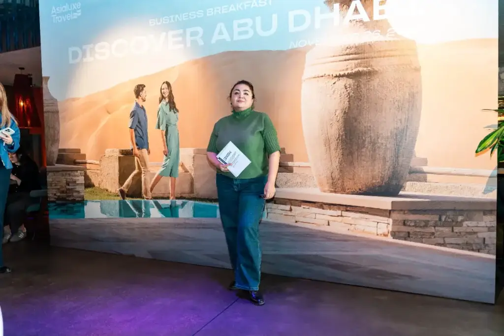 Женщина в зелёной одежде стоит перед постером бизнес-завтрака „Discover Abu Dhabi“, держа в руках буклет с информацией о мероприятии