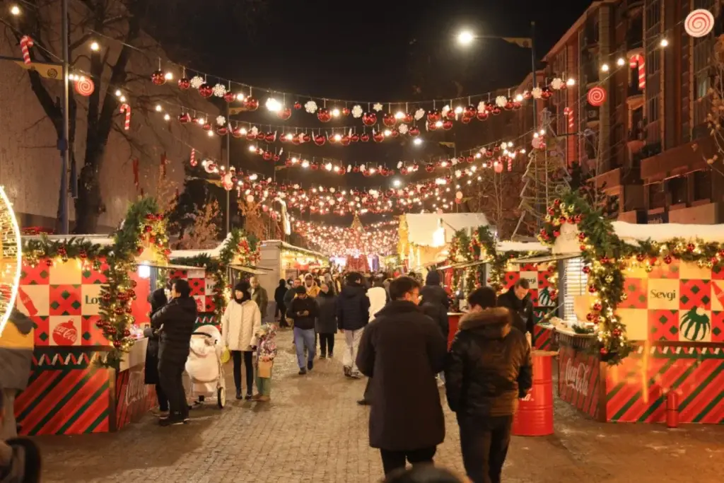 A festive Christmas market at night with illuminated stalls, holiday garlands, hanging ornaments, and crowds of people walking along a decorated street.