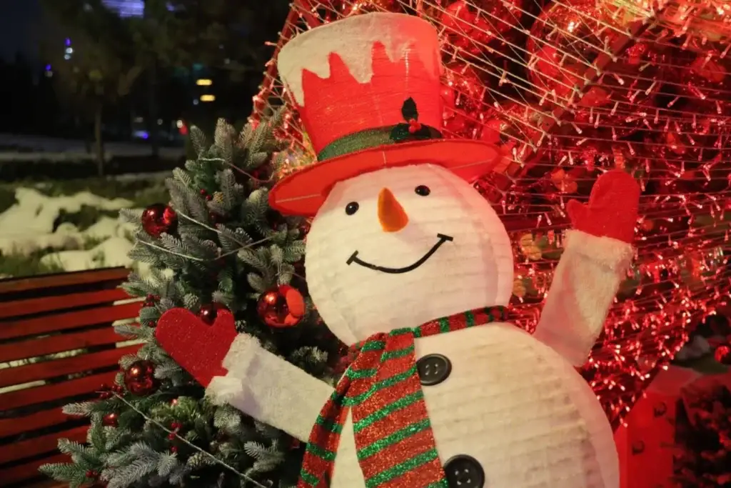 A cute smiling snowman with a red hat, wearing a red and green scarf, against a backdrop of Christmas decorations