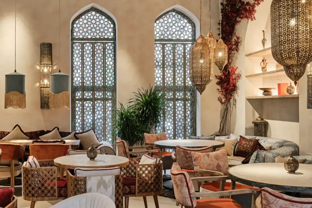 Интерьер восточного ресторана: арочные окна с резными решётками, подвесные светильники, мягкие диваны с подушками, круглые столы, декоративные керамические сосуды, зелёные растения, полки с восточными аксессуарами