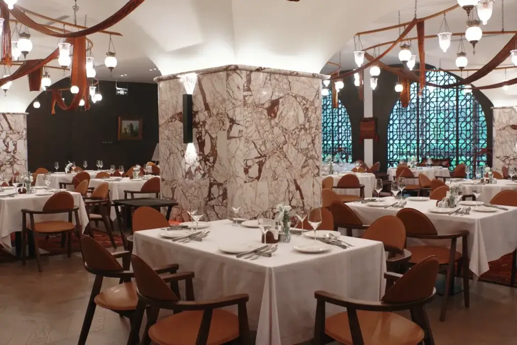 Ресторанный зал с мраморными колоннами, подвесными светильниками и сервированными столами, накрытыми белыми скатертями. Коричневые стулья дополняют изысканный интерьер