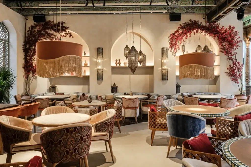Интерьер восточного ресторана: арочные окна с резными решётками, подвесные светильники, мягкие диваны с подушками, круглые столы, декоративные керамические сосуды, зелёные растения, полки с восточными аксессуарами