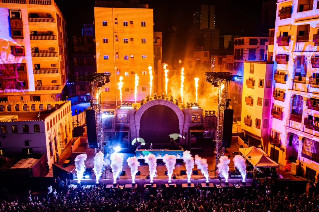 Night outdoor concert in an urban setting with a large stage, fire and smoke effects, colorful lighting, and a crowd gathered in front of surrounding buildings