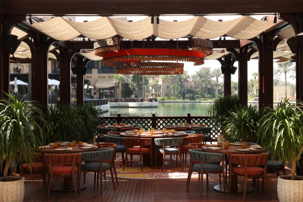 Open-air restaurant terrace with set tables under a canopy by a water feature and palm trees