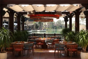 Open-air restaurant terrace with set tables under a canopy by a water feature and palm trees