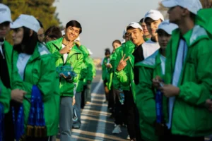 A group of young people in green windbreakers and white caps posing on the street, some showing victory signs