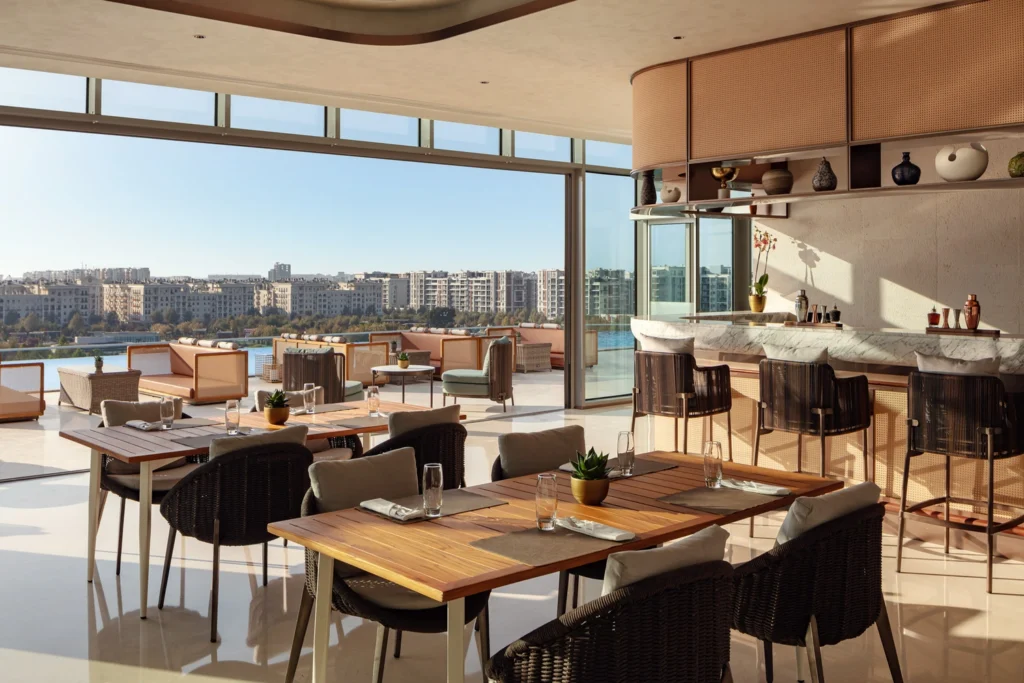 An outdoor restaurant or bar terrace with wooden tables, woven chairs, and a panoramic view of a cityscape and water
