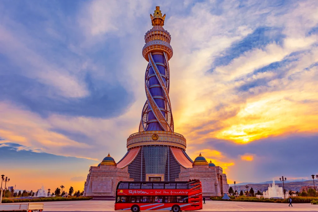 The Istiklol Monument in Dushanbe at sunset, a tall golden tower with a crown on top, surrounded by fountains and a city square with a red sightseeing bus in the foreground