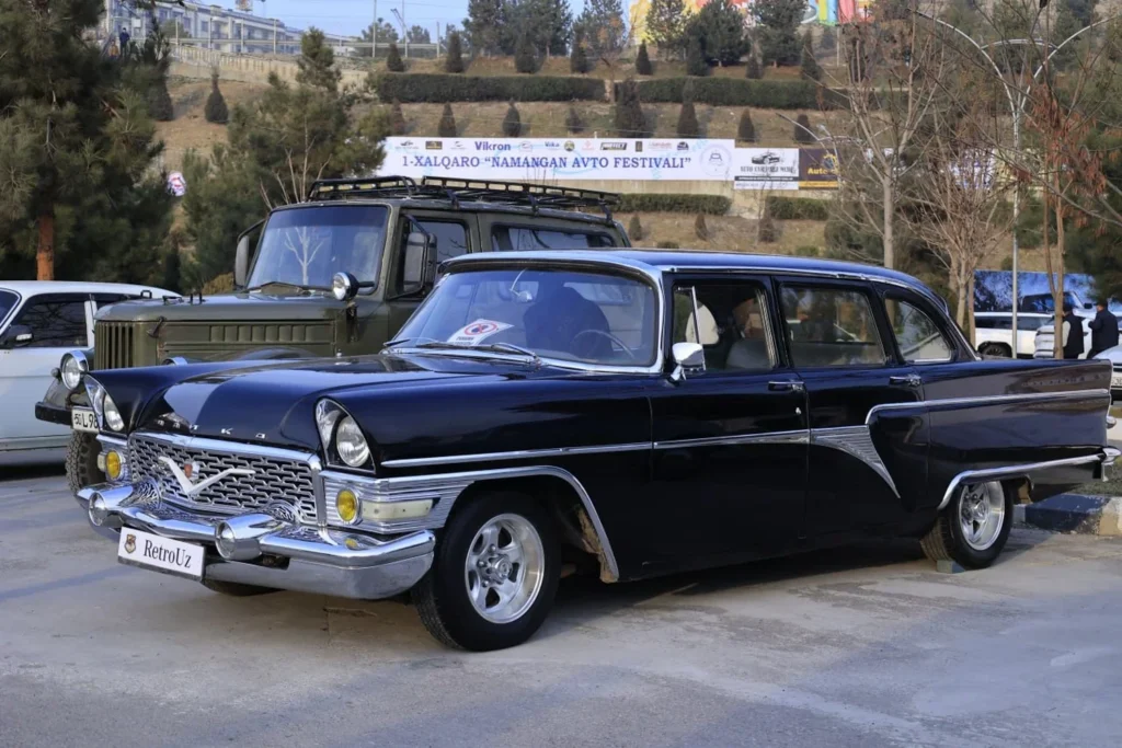 A black classic GAZ-13 "Chaika" car with chrome bumpers and a grille, parked next to a green military GAZ-66 truck at the "NAMANGAN AVTO FESTIVALI" car festival