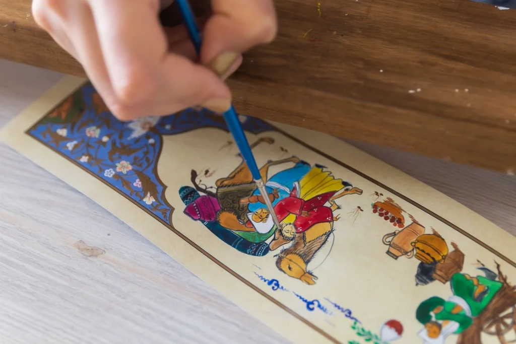 Artist hand painting a miniature illustration with a fine brush, depicting figures and a camel in a traditional oriental style
