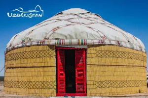 A traditional yurt with a white dome, reed walls, and a bright red wooden door against a clear blue sky with the “Uzbekistan” logo in the corner