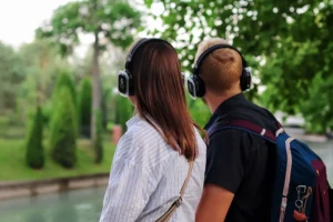 A guy and a girl stand with their backs to the camera, wearing headphones, and look at the river ahead and the fir trees on the green lawn