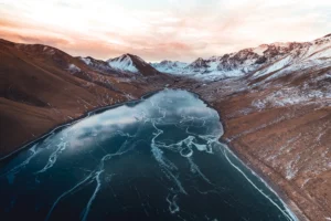 A high-altitude frozen lake named Kol-Ukok, surrounded by mountains with patches of snow under an evening sky, in Kyrgyzstan