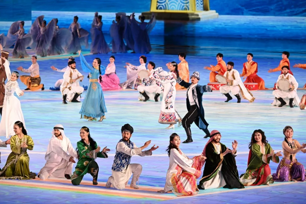 A cultural performance likely at the 2025 Harbin Asian Winter Games opening ceremony, featuring dancers in colorful traditional Uzbek and other Asian costumes on a large stage with a snow