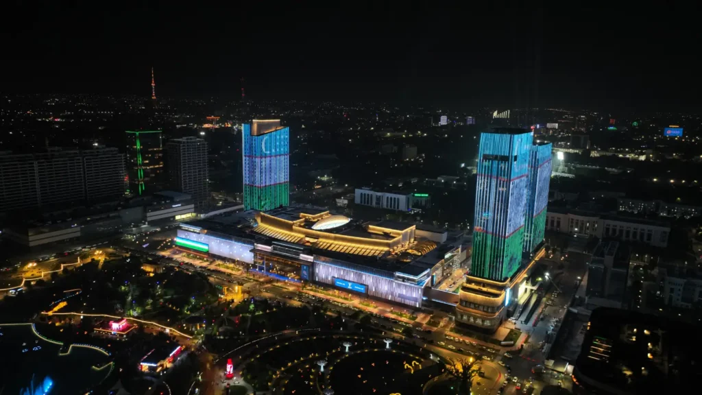 Night aerial view of Tashkent City Mall illuminated with lights, surrounded by city buildings