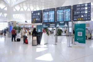 На фотографии показан интерьер терминала Международного аэропорта имени короля Халида в Эр-Рияде, Саудовская Аравия. Видны пассажиры с багажом, стойка регистрации или информационный киоск, а также большие электронные табло с информацией о вылетах, указателями на выходы к самолетам, туалеты и молельную комнату