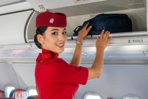Стюардесса в красной форме кладет синюю сумку в верхний отсек самолета