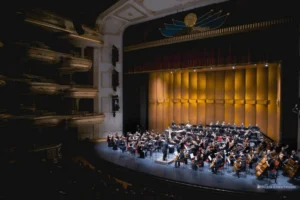 An orchestra performs on the stage of a luxurious opera house or concert hall, featuring balconies, yellow curtains, and hieroglyphic ornament above the stage